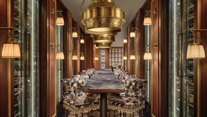 Une longue et élégante table à manger, agrémentée de chaises rembourrées, est flanquée de vitrines. L’ensemble se trouve dans un couloir richement décoré, illuminé par un grand et somptueux lustre.