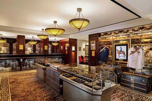 Un hall d’entrée luxueux, agrémenté de lustres somptueux, où sont exposés des tenues de soirée élégantes, et dont le sol est recouvert d’un tapis à motifs.
