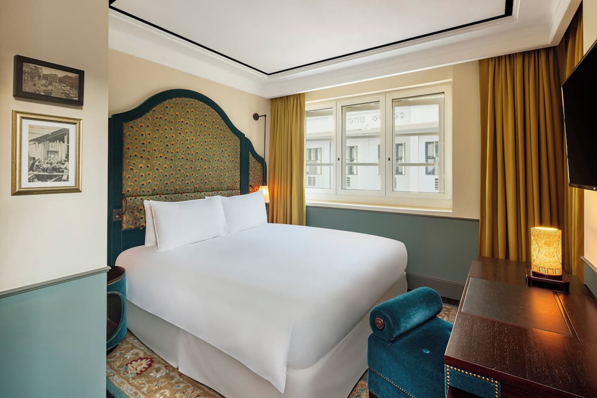 Ein Hotelzimmer mit Bett, Schreibtisch, Stuhl, Lampe, einem Fenster mit Vorhängen und gerahmten Bildern an der Wand.