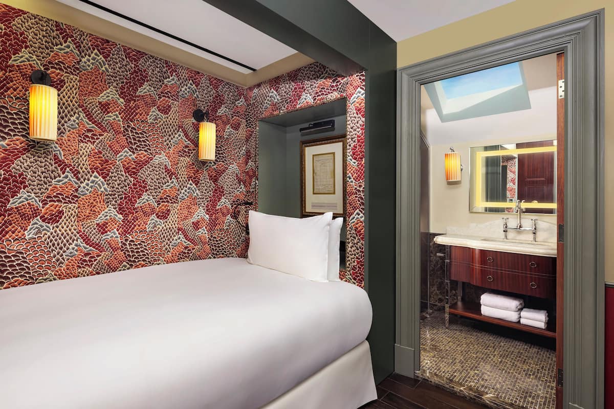 Ein Hotelzimmer mit Wandmuster, einem Bett, einem Badezimmer mit Waschbecken und Spiegel sowie einem gerahmten Bild an der Wand.