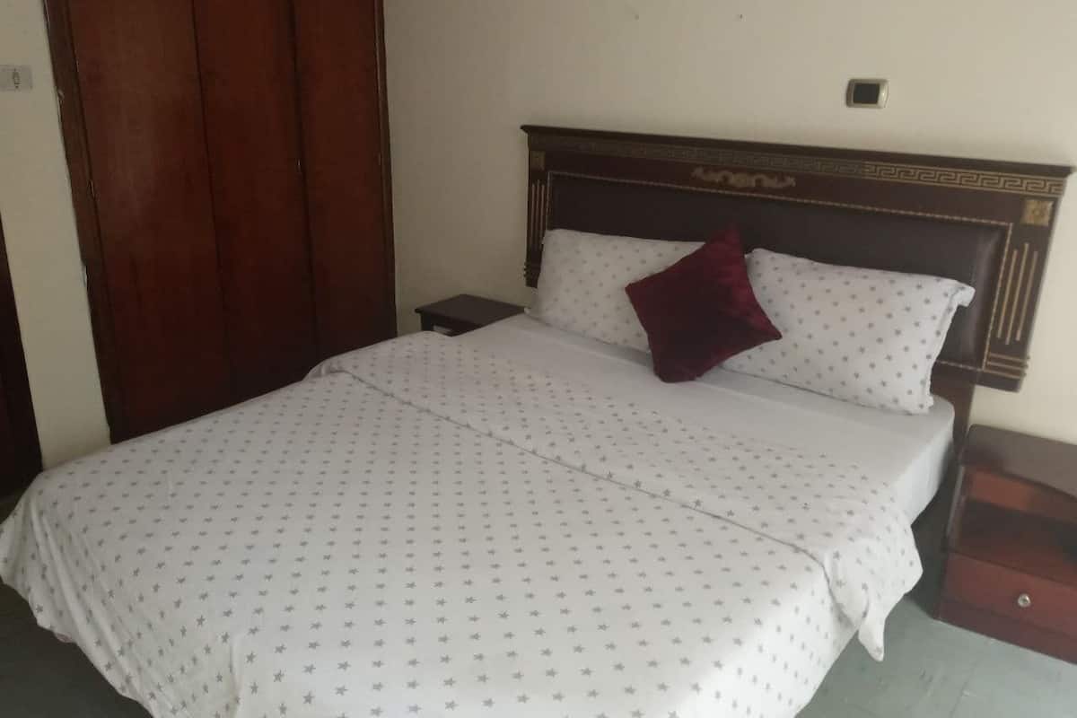 A bed with a white bedspread and a red pillow, a wooden headboard, a small bedside table, and a wooden wardrobe.