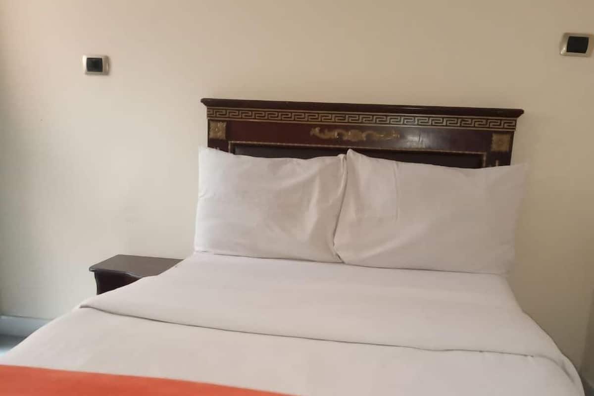 A bed with white linens and an orange blanket, a wooden headboard with a decorative pattern, and a small nightstand with a lamp.