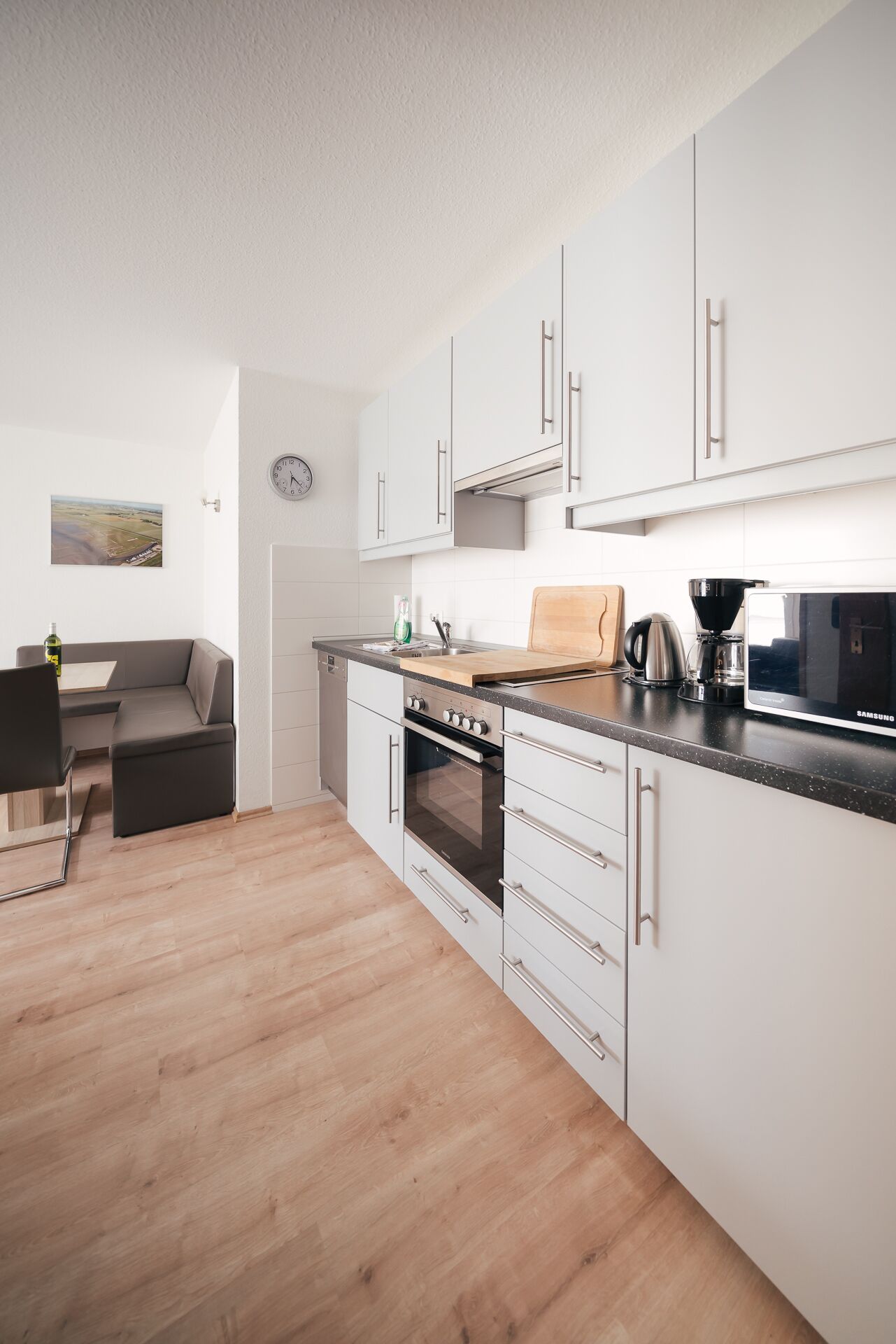 A modern kitchen with white cabinets, a built-in oven, and a microwave. There is a wooden cutting board on the counter and a coffee maker.