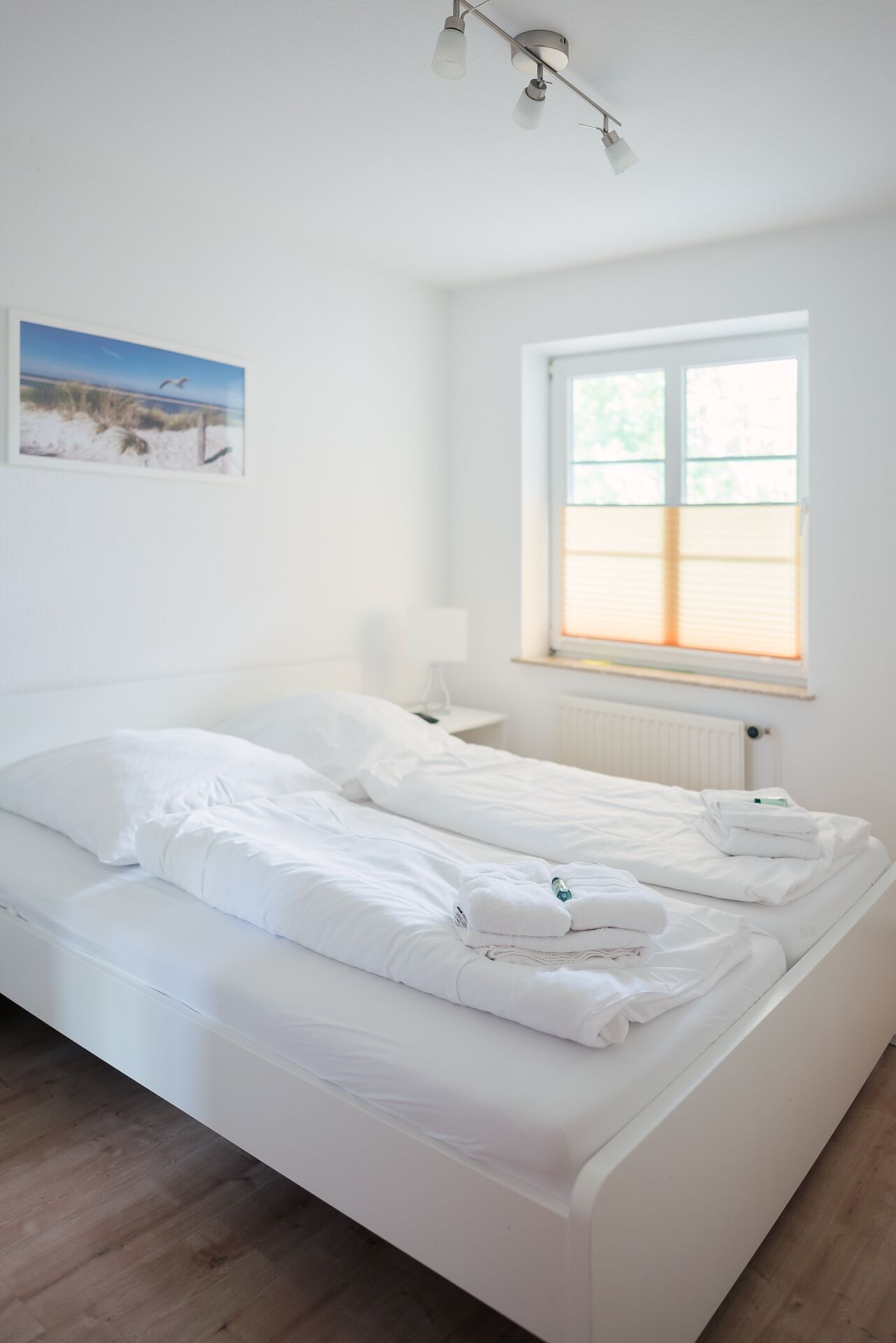 Ein sauberes, weißes Schlafzimmer mit einem Bett, einem Fenster mit Jalousien, einer Nachttischlampe und einem gerahmten Bild an der Wand.