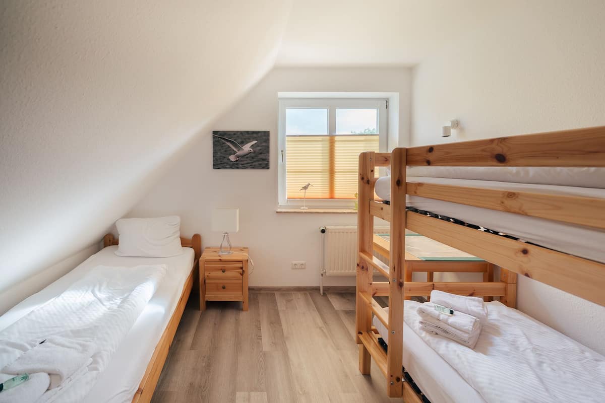 Ein kleiner, gut beleuchteter Raum mit zwei Etagenbetten, einem Einzelbett, einem Nachttisch, einer Lampe und einem Fenster mit Jalousien.
