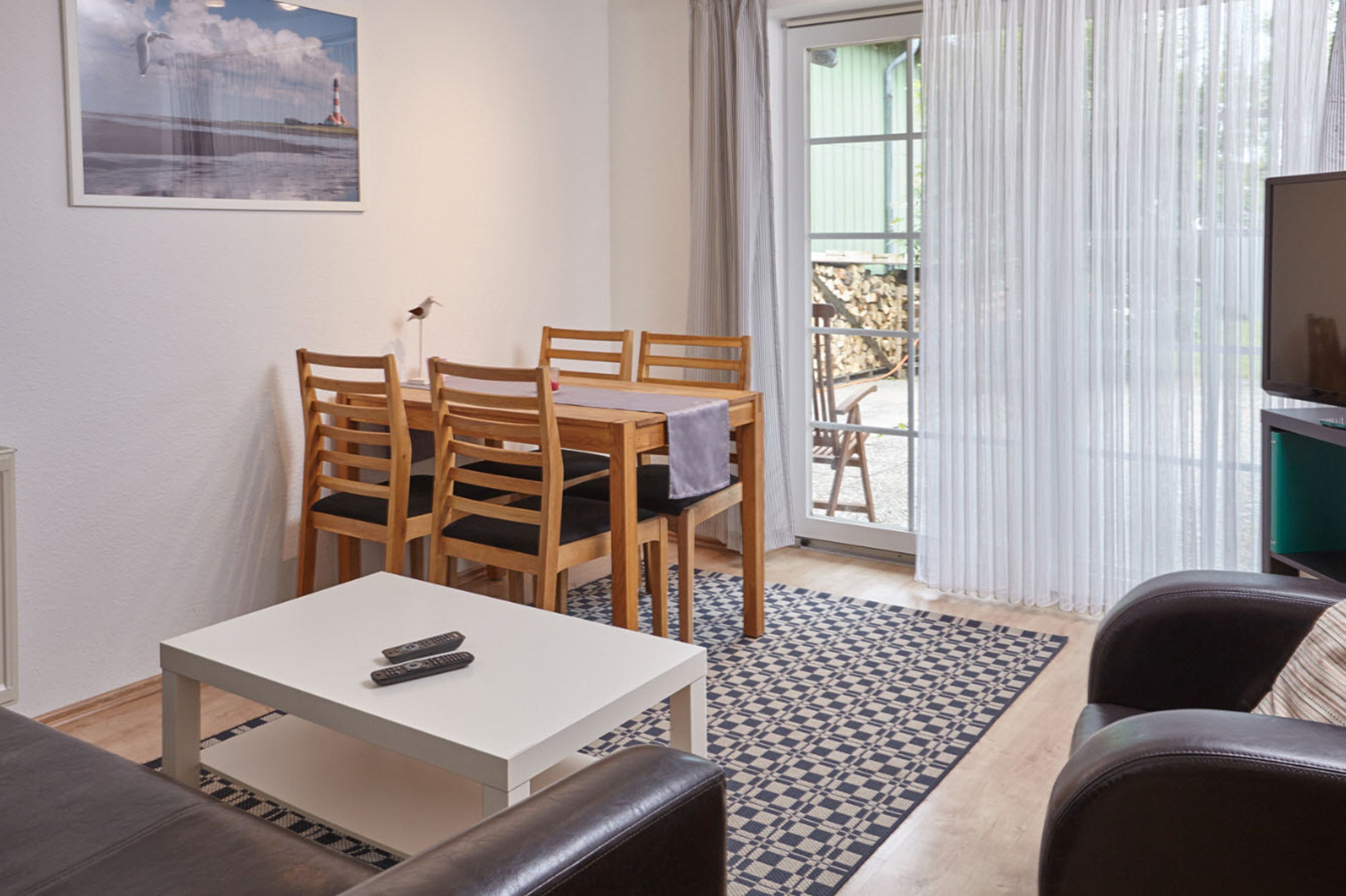 Quarto com mesa de jantar e cadeiras, um sofá, uma TV e vista para o exterior através de uma janela com cortinas leves.