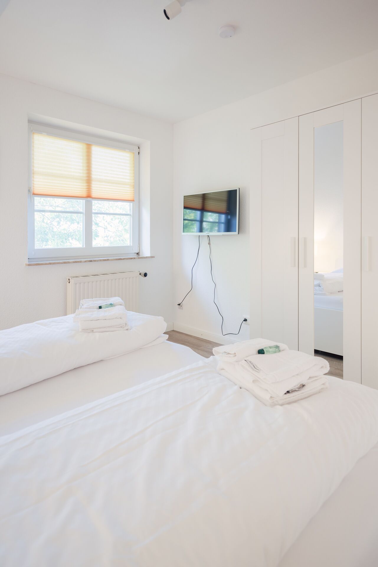 A hotel room with two beds, a window with blinds, a radiator, a flat-screen TV mounted on the wall, and a wardrobe.