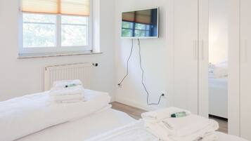 A hotel room with two beds, a window with blinds, a radiator, a flat-screen TV mounted on the wall, and a wardrobe.