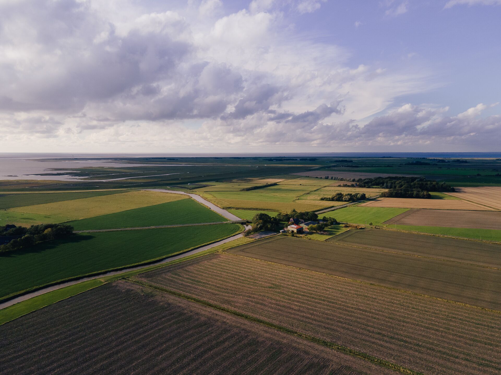An aerial view of a rural landscape with fields, a small cluster of buildings, and a body of water in the distance.