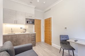 A modern kitchen with grey cabinets, a built-in oven, and a coffee maker. There is a wooden door and a small table with a chair. - Alexander Apartments (London)