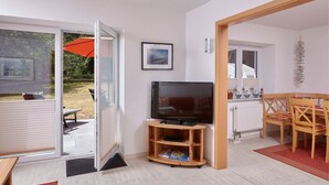Una sala de estar con televisión, un mueble bajo de madera para la televisión, una mesa de comedor con sillas y un ventanal corredero que da a un espacio exterior con sombrilla.