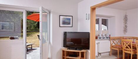 A living room with a television, a wooden TV stand, a dining table with chairs, and a sliding glass door leading to an outdoor area with an umbrella.