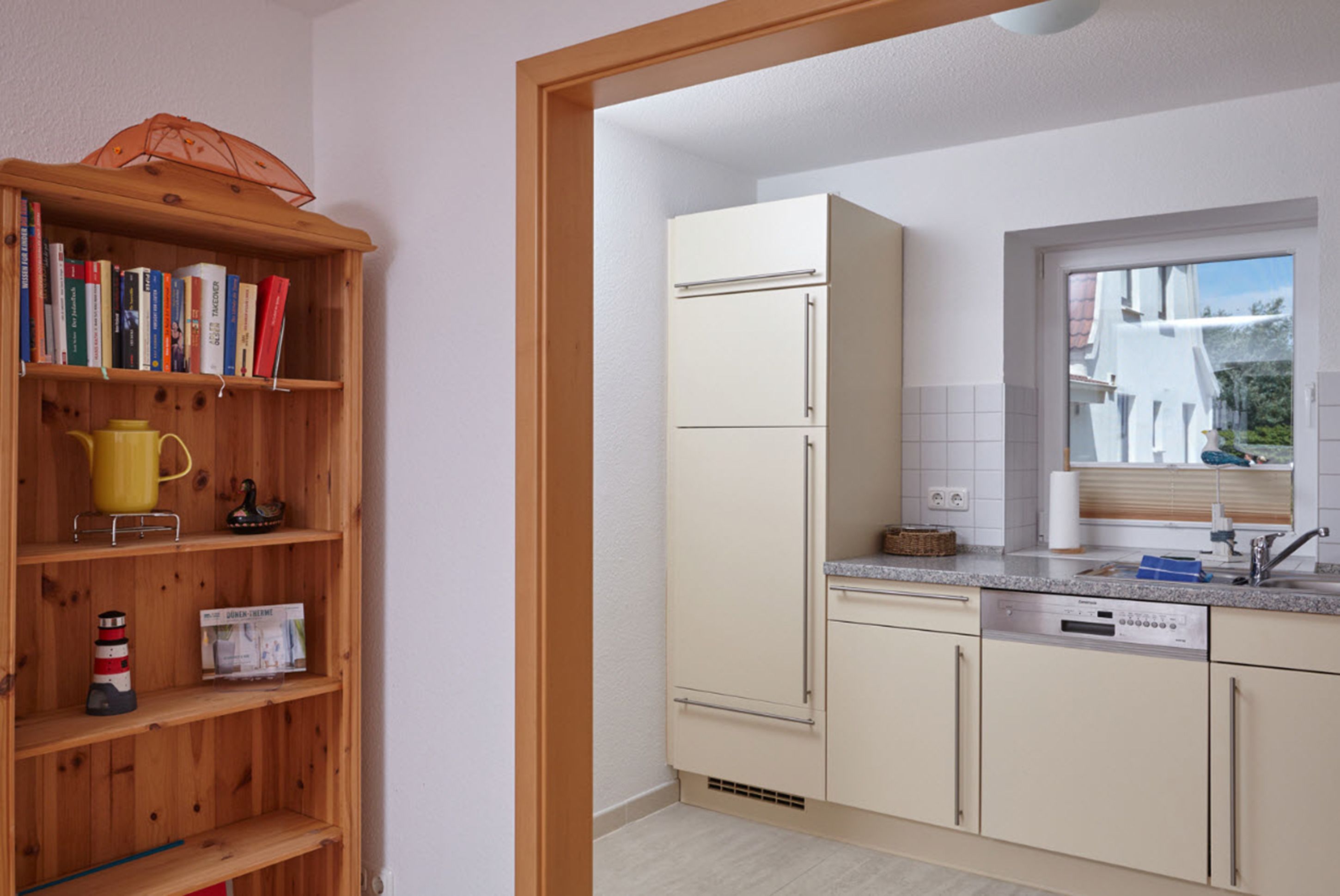 A kitchen with a wooden bookshelf, a refrigerator, a dishwasher, and a window with a view of buildings.