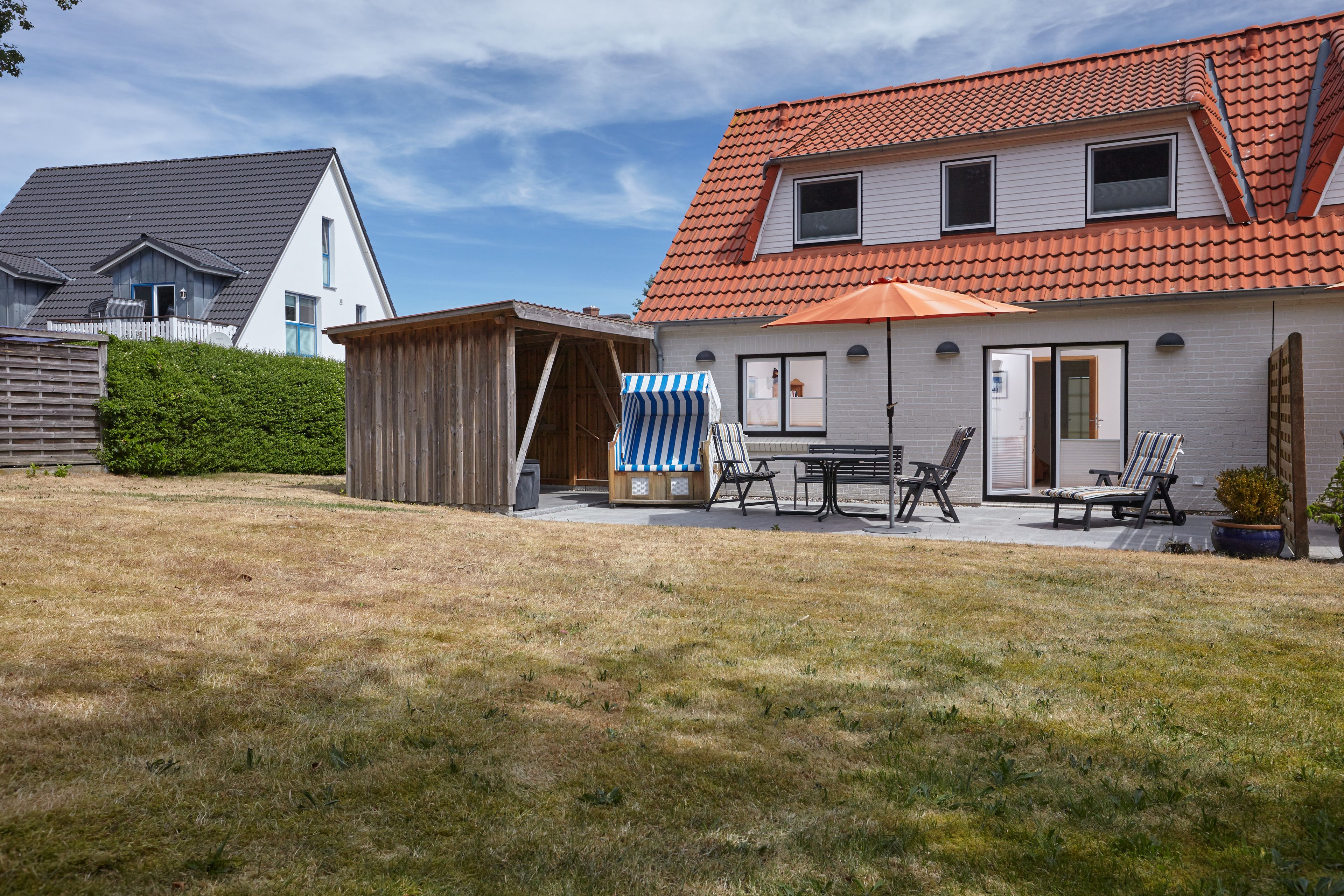 Une maison à deux étages avec un toit recouvert de tuiles rouges, une cour clôturée et une terrasse aménagée avec du mobilier d’extérieur.
