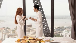 A bed with breakfast items, a laptop, and a view of a cityscape through large windows. - The Snowbell Hotel & SPA @ The Bridge. (Phnom Penh)