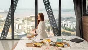A bed with breakfast tray, a laptop, and a view of a cityscape through large windows. - The Snowbell Hotel & SPA @ The Bridge. (Phnom Penh)