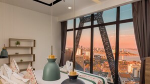 A modern bedroom with a large window, a bed with floral bedding, a bedside table, and a hanging light fixture. - The Snowbell Hotel & SPA @ The Bridge. (Phnom Penh)