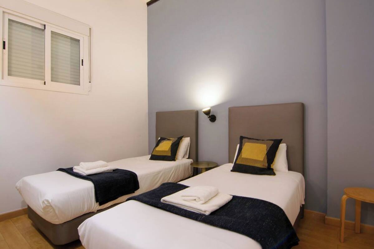 A hotel room with two beds, each with a headboard, a bedside table, and a wall-mounted lamp.