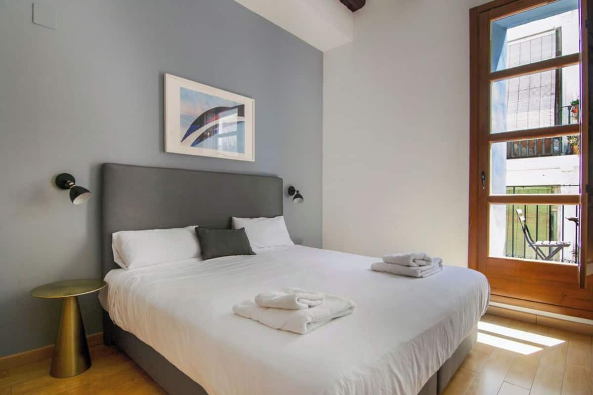 A bedroom with a bed, a round side table, a framed picture on the wall, and a window with a view of the outside.