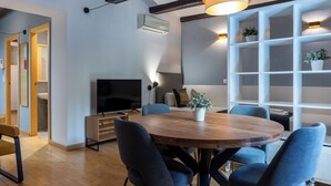 A modern living room with a wooden dining table, blue chairs, a TV, and a white shelving unit with plants. - Welcomer Apartments Valencia (Valencia)