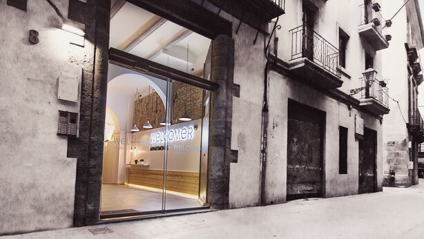 A modern apartment building with a glass entrance, a visible interior with a reception desk, and a balcony with railings. - Welcomer Apartments Valencia (Valencia)