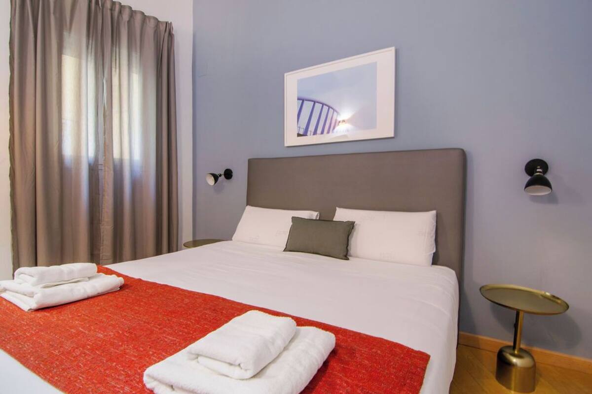A hotel room with a bed, a red throw blanket, white towels, a framed picture on the wall, a small table, and a lamp.