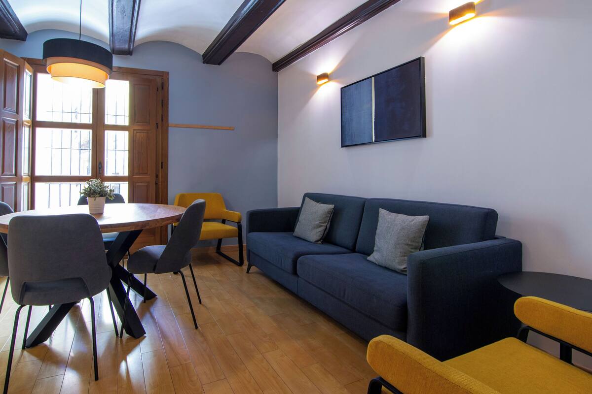 A modern living room with a dark blue sofa, yellow chairs, a wooden dining table, and a large window with natural light.