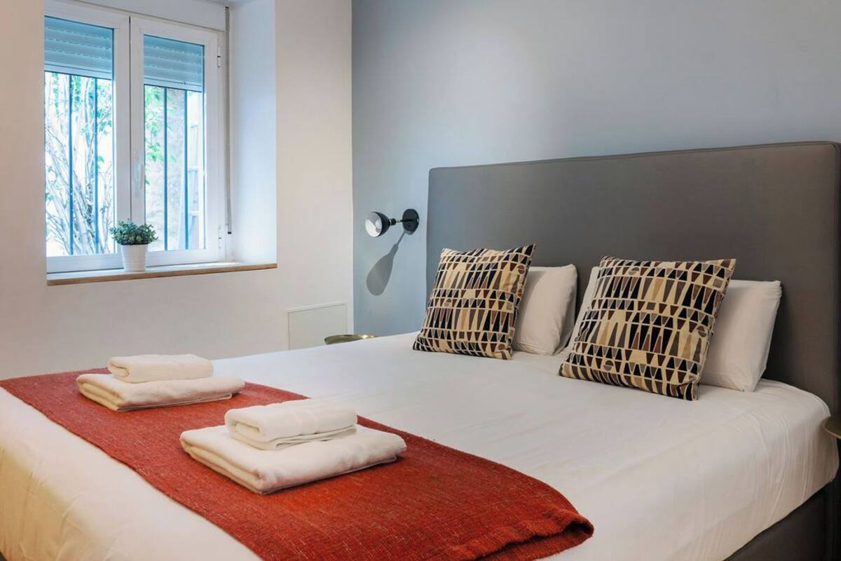 A neatly made bed with white linens and a red throw blanket, two patterned pillows, a bedside lamp, and a window with blinds.