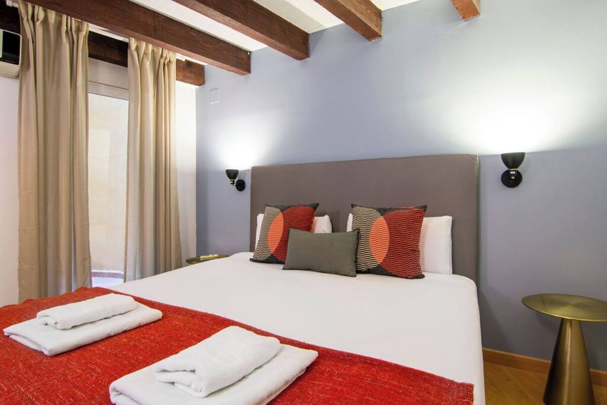 A hotel room with a large bed, beige curtains, a red throw blanket, and a round gold side table.