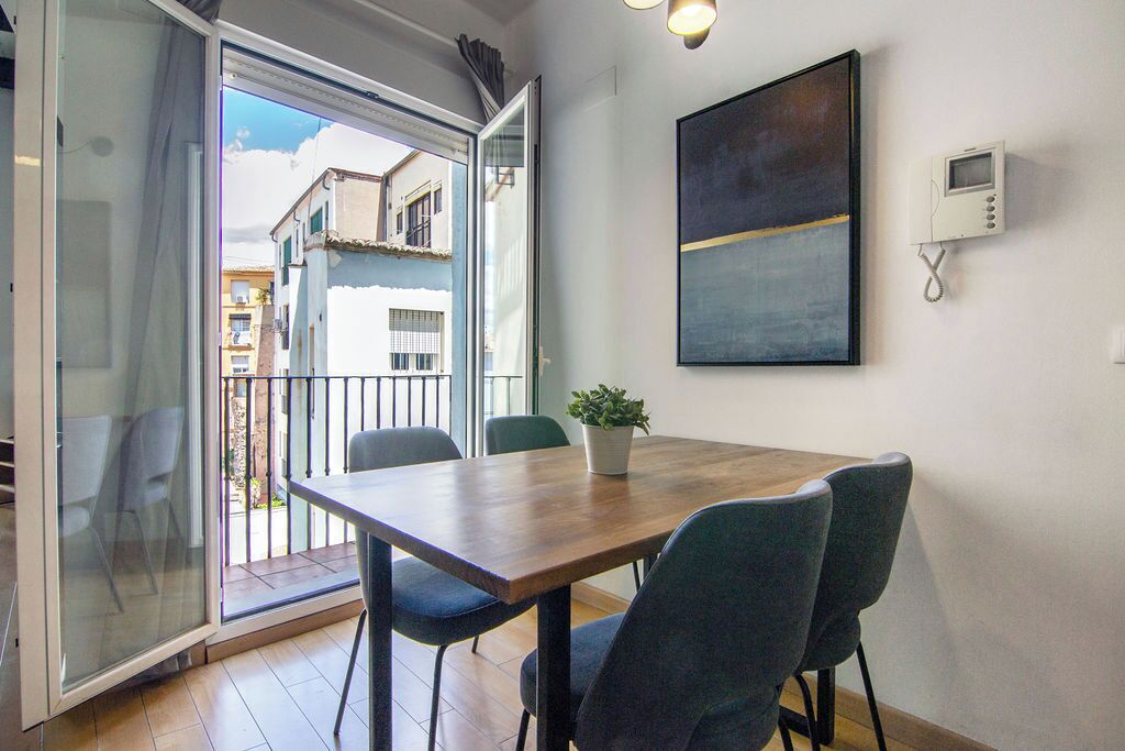 A dining area with a wooden table, four chairs, a potted plant, and a view of buildings through a glass door.