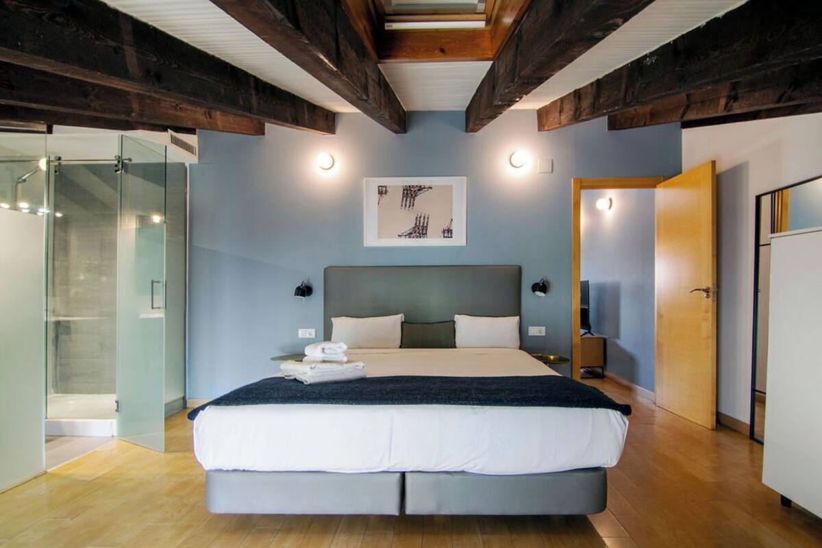 A modern bedroom with a large bed, a glass shower enclosure, and a wooden ceiling.