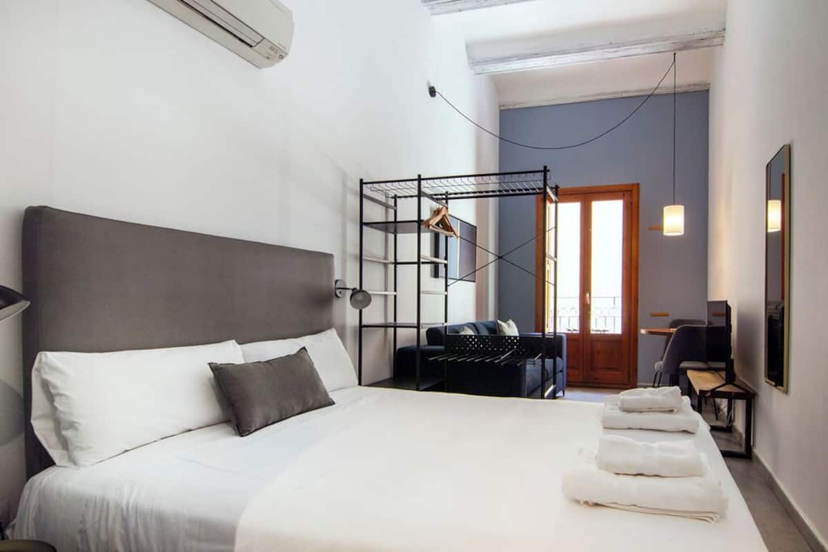 A hotel room with a large bed, a desk, a chair, a wardrobe, and a door leading to a balcony.