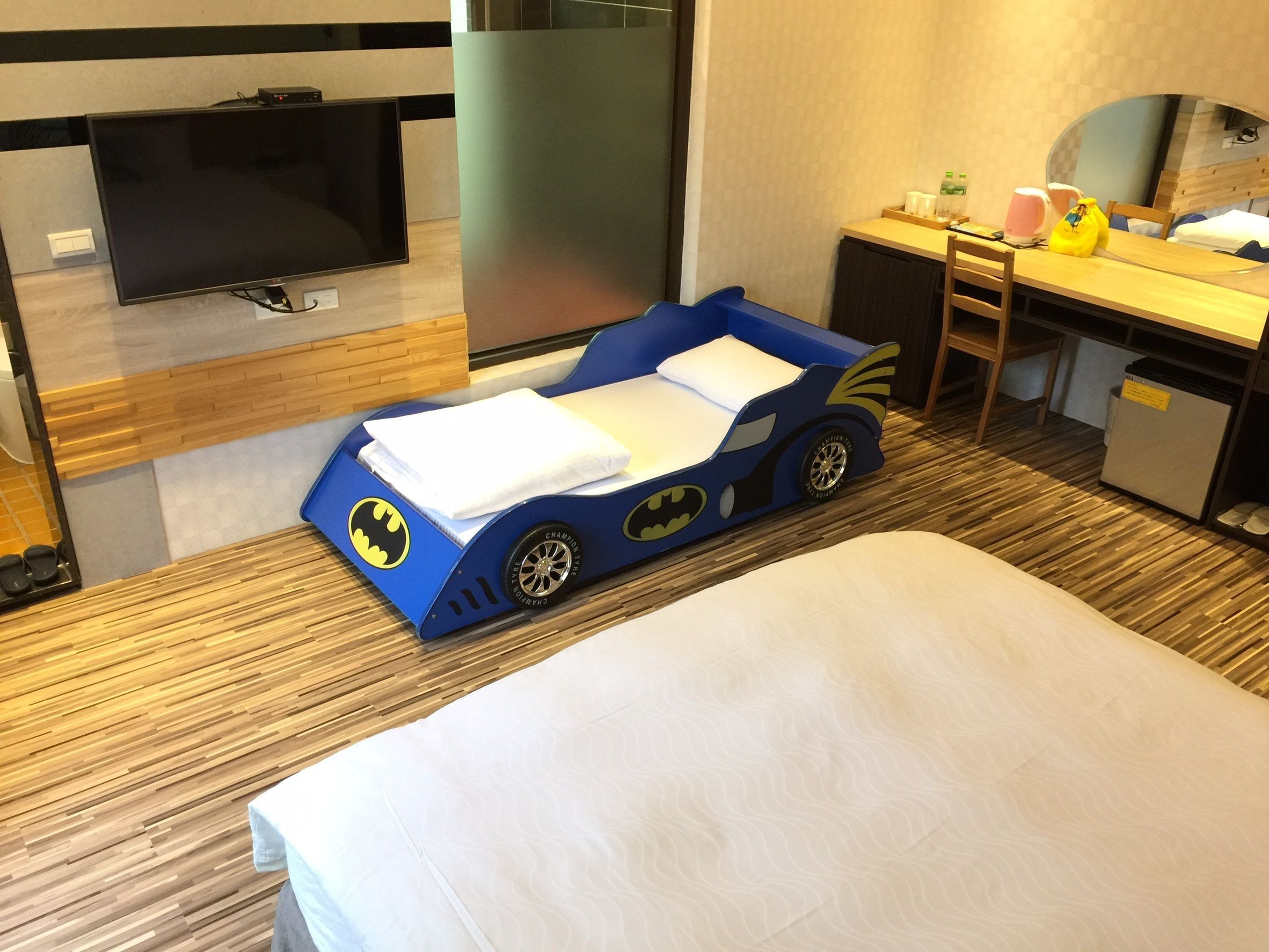 Una habitación de hotel con una cama, un escritorio, una televisión y un coche de juguete con temática de Batman.