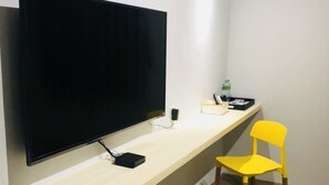 A room with a large flat-screen TV mounted on a wall, a wooden desk with a chair, and a small table with a lamp and a bottle. - Ho Young Hotspring Hotel (Jiaoxi)