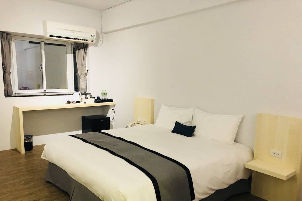 A hotel room with a bed, bedside tables, a desk with a chair, an air conditioning unit, and a window with curtains.