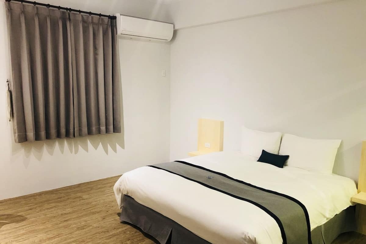 A hotel room with a bed, curtains, an air conditioning unit, a bedside table with a lamp, and a wooden floor.