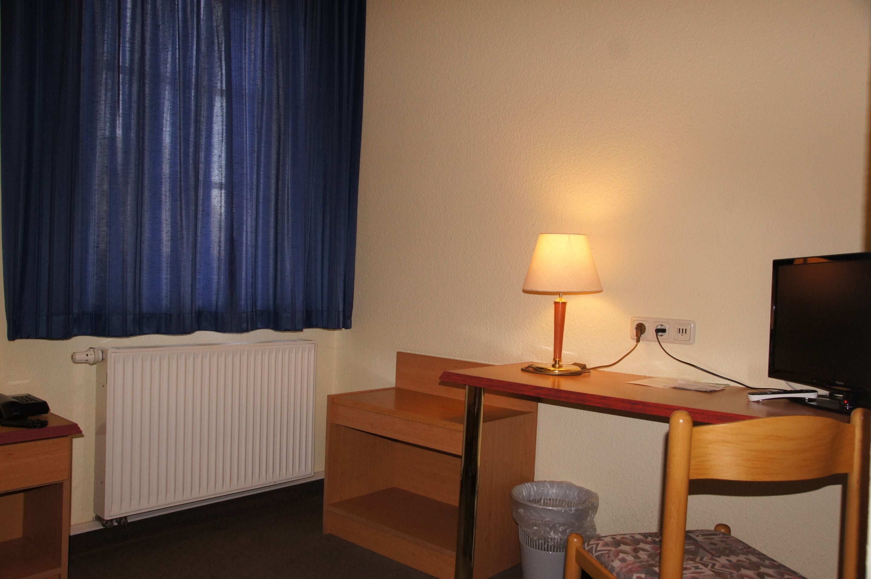 Une petite pièce bien rangée, avec un bureau en bois, une lampe, une chaise, un radiateur et un téléviseur.