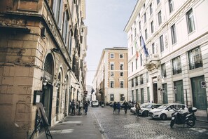 A cobblestone street lined with historic buildings, a flag, and parked cars. - Spagna Rooms (Rome)