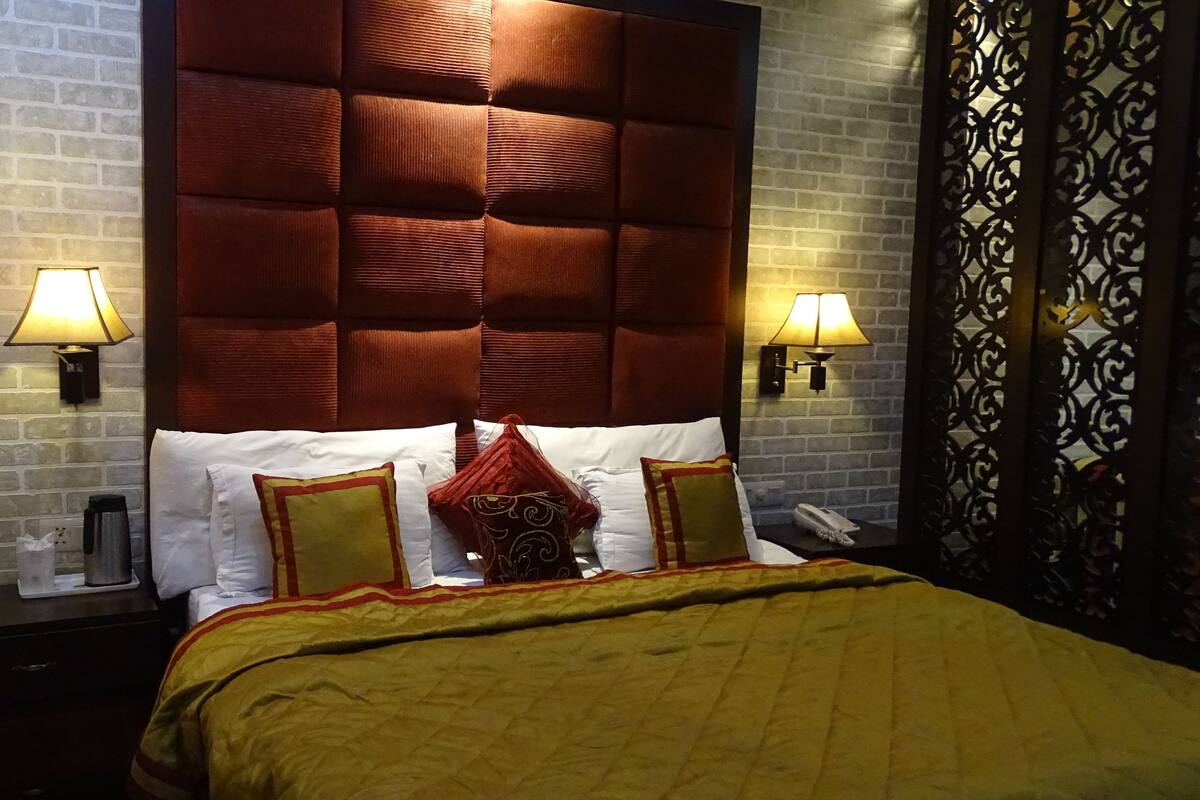A hotel room with a brick wall, a tufted headboard, a bed with white and red pillows, and two bedside lamps.