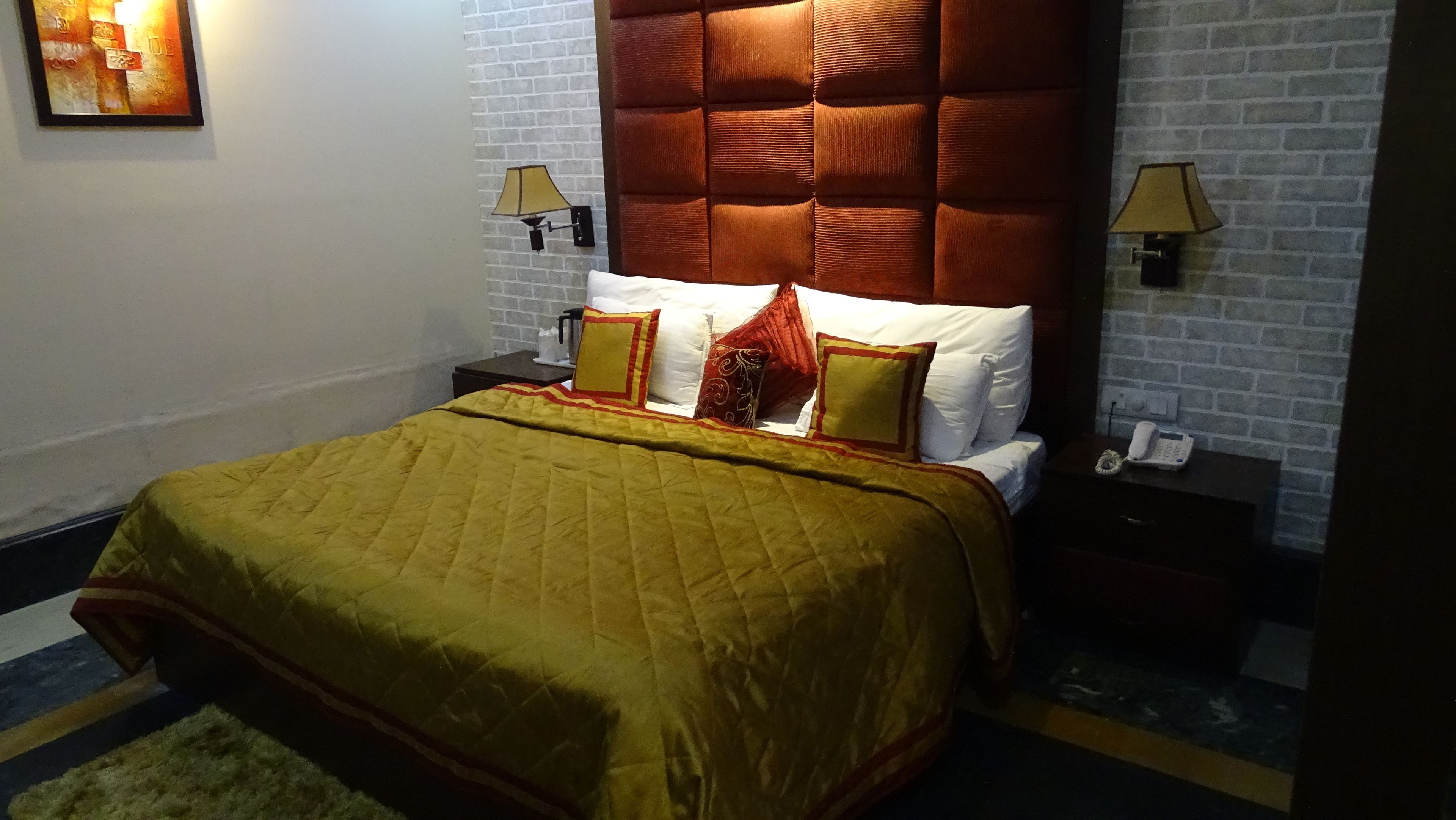 A hotel room with a brick wall, a tufted headboard, a bed with a yellow quilt, and two bedside lamps.