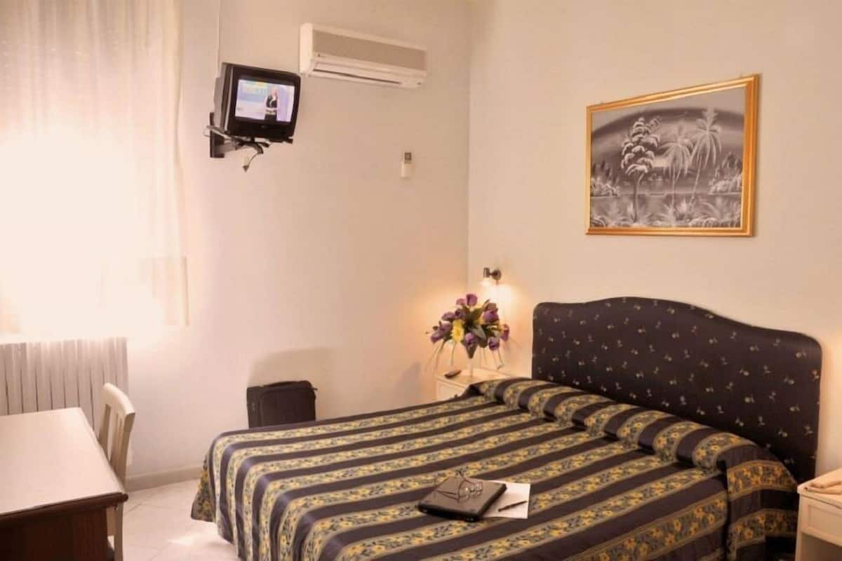 A hotel room with a bed, a TV mounted on the wall, a framed picture, and a desk with a chair.