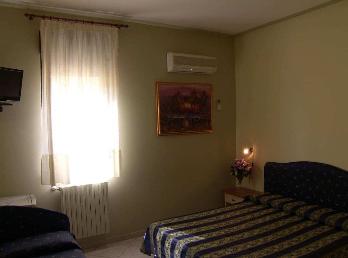 A bedroom with a bed, a nightstand with a lamp, a painting on the wall, and a window with curtains.