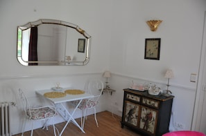 Una habitación con una mesa blanca, dos sillas, un espejo, un armario con pinturas y un radiador.