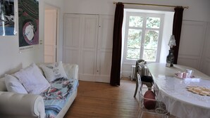 A room with a white sofa, a dining table, a chair, a radiator, a window with curtains, and a poster on the wall.