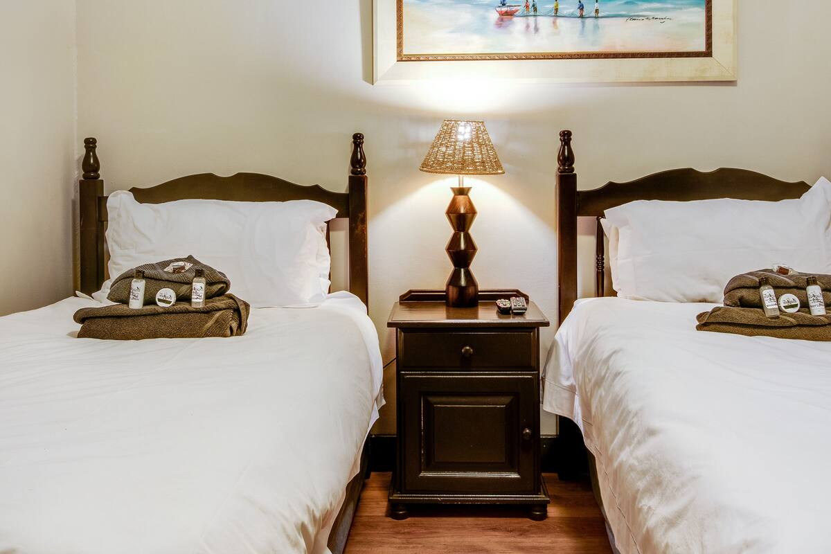 A hotel room with two beds, a nightstand with a lamp, and a painting of a beach scene on the wall.