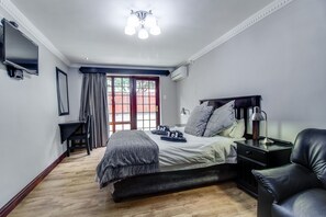A bedroom with a bed, bedside table, lamp, and a TV mounted on the wall. - The Nightingale Guesthouse (Bloemfontein)