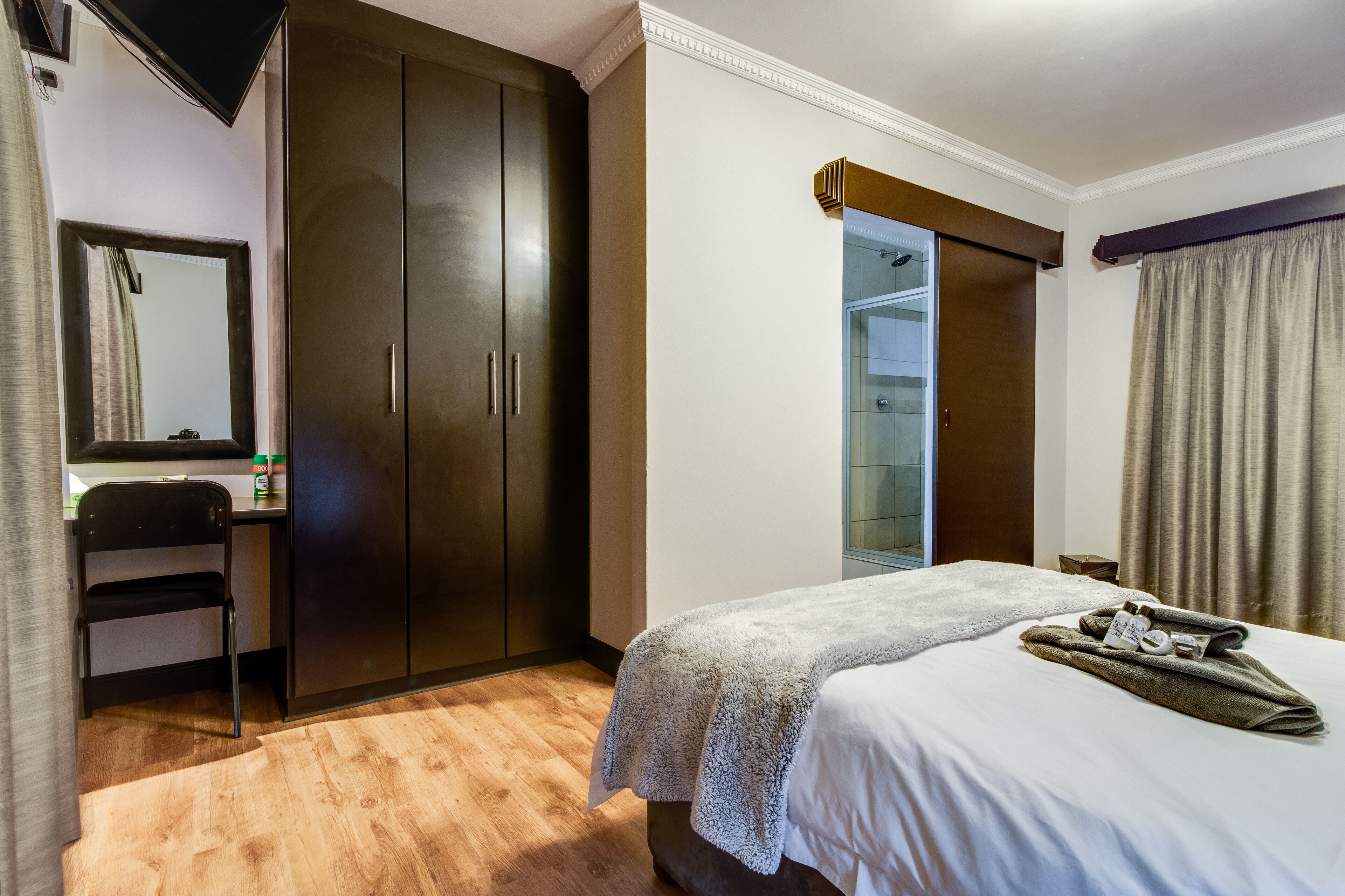 A hotel room with a bed, a wardrobe, a desk with a chair, a mirror, and a bathroom visible through a glass door.
