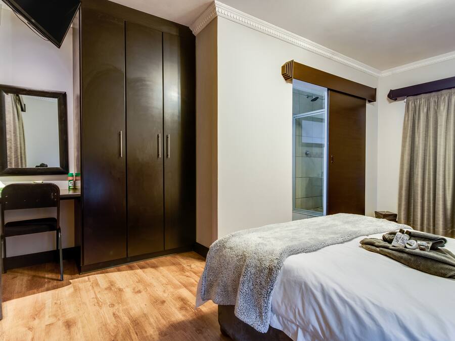A hotel room with a bed, a wardrobe, a desk with a chair, a mirror, and a bathroom visible through a glass door.