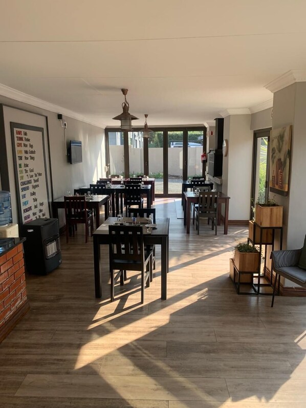 A spacious dining area with multiple tables and chairs, large windows, and a ceiling fan. - The Nightingale Guesthouse (Bloemfontein)