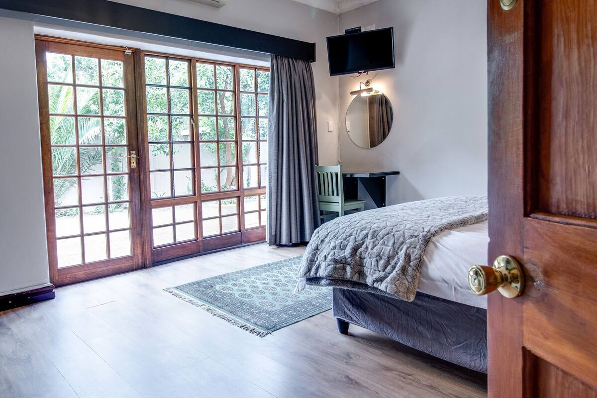 A bedroom with a bed, a desk, a chair, a TV, and a large window with a view of greenery.